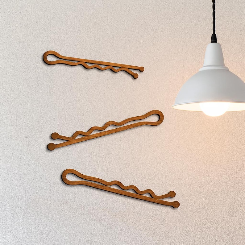 May include: Three wooden bobby pins in a warm brown hue are arranged on a textured white wall. The pins have a classic wavy design and are evenly spaced. A white pendant light hangs in the background.