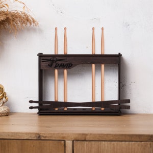 Pode incluir: Um suporte para baquetas de madeira marrom escuro com o nome personalizado "DAVID" gravado na frente. O suporte tem ranhuras para guardar as baquetas e um design em forma de cruz com um punho segurando as baquetas.