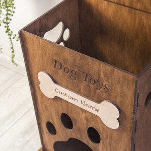 May include: A wooden dog toy storage bin with paw-shaped cutouts and a bone-shaped nameplate. The words "Dog Toys" are engraved on the front. The bin is made of dark brown wood.