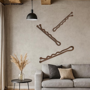 May include: Three large, chestnut-colored bobby pins are mounted on a textured wall. The bobby pins are arranged in a diagonal pattern. The wall decor is above a gray sofa with decorative pillows. A black pendant light hangs from the ceiling.