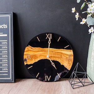 May include: A black and gold clock with Roman numeral markings. The clock face is made of epoxy resin with a wood inlay. The clock is 12 inches in diameter.