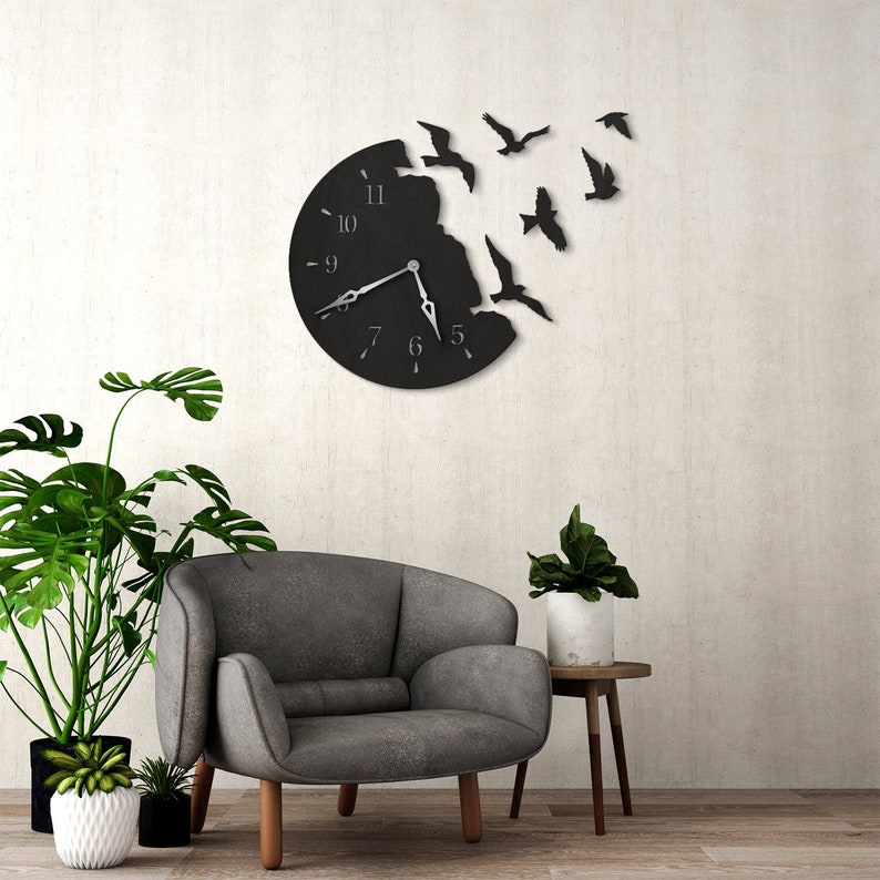 May include: Black wall clock with a broken circle design and birds flying out of the opening. The clock has silver hands and black Roman numeral markings. The clock is hanging on a white wall with a gray armchair and a potted plant in front of it.