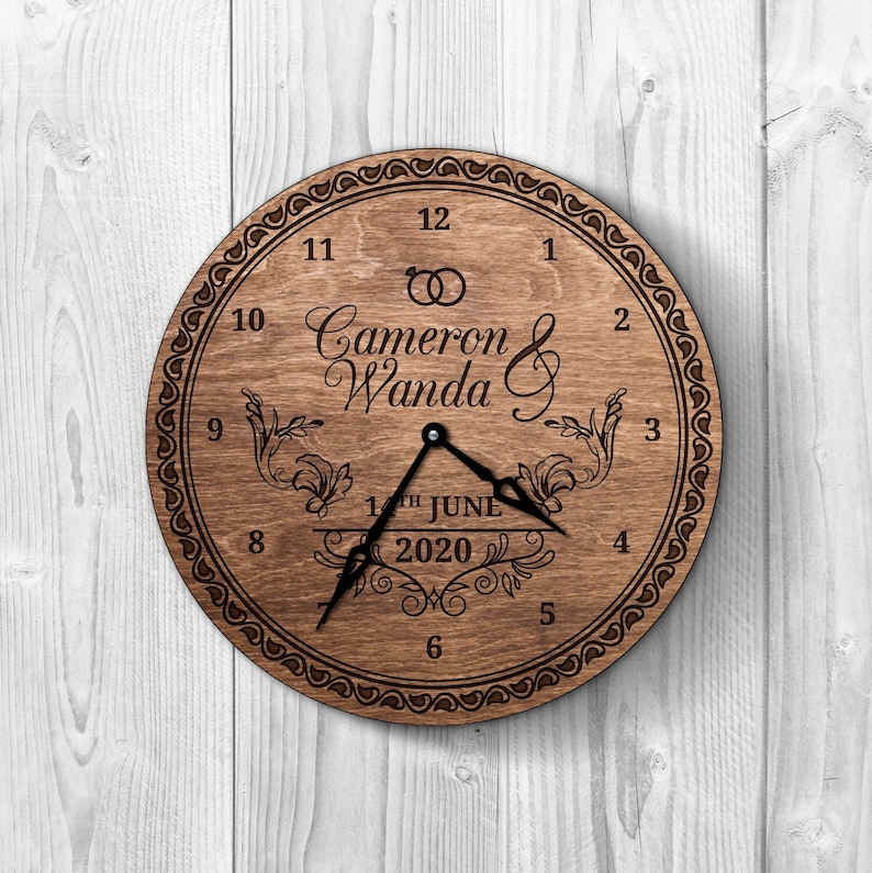 May include: A wooden clock with a brown background and black hands. The clock face has a decorative border and the text "Cameron & Wanda" with a wedding ring symbol above the names. The date "12th June 2020" is written below the names.