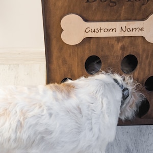 May include: A wooden dog toy box with a bone-shaped cutout that reads "Custom Name." The box has circular holes and a dog is interacting with it. The dog is white and tan.