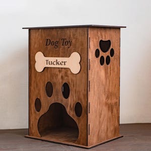 May include: A wooden dog toy box with a paw print design and the name "Tucker" on a bone-shaped plaque. The box is brown with the words "Dog Toy" engraved on the front. The box has a paw print cut out on the side.