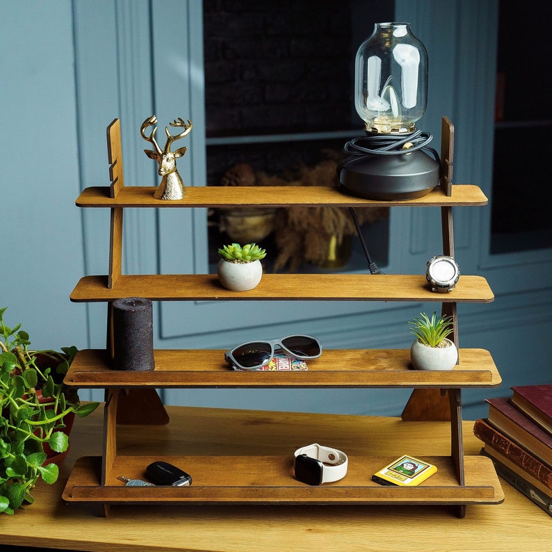 May include: A wooden tiered shelf unit with four shelves. The top shelf has a gold deer figurine and a black lamp with a clear glass shade. The second shelf has a small potted plant. The third shelf has sunglasses and a potted plant. The bottom shelf has a watch, keys, and a small box.