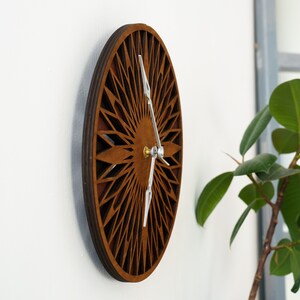K&ouml;nnte beinhalten: Eine braune Wanduhr aus Holz mit geometrischem Design. Die Uhr hat silberne Zeiger und eine Messingmitte.