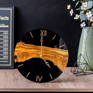 May include: A black and gold clock with Roman numeral markings. The clock face is made of epoxy resin and features a natural wood inlay.
