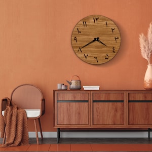 May include: A wooden clock with Arabic numerals on a brown wall. The clock is hanging above a wooden cabinet with three drawers. There is a chair and a blanket in the foreground.