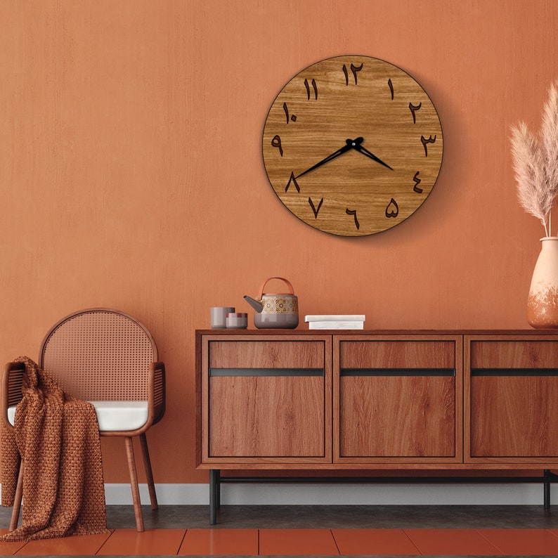May include: A wooden clock with Arabic numerals on a brown wall. The clock is hanging above a wooden cabinet with three drawers. There is a chair and a blanket in the foreground.