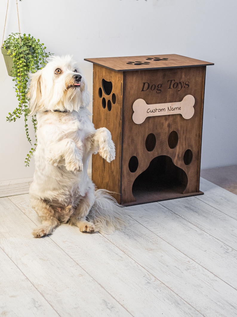 May include: A wooden dog toy box with paw print cutouts and the words "Dog Toys" and "Custom Name". A white dog stands on its hind legs next to the box. A hanging plant is in the background.