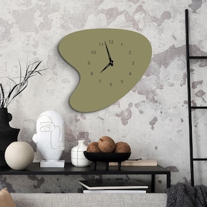 May include: A green, abstractly shaped wall clock with black hands and numbers. The clock is hanging on a textured, light gray wall. The clock is surrounded by a black shelf with a white sculpture, a white vase, and a black bowl with coconuts.