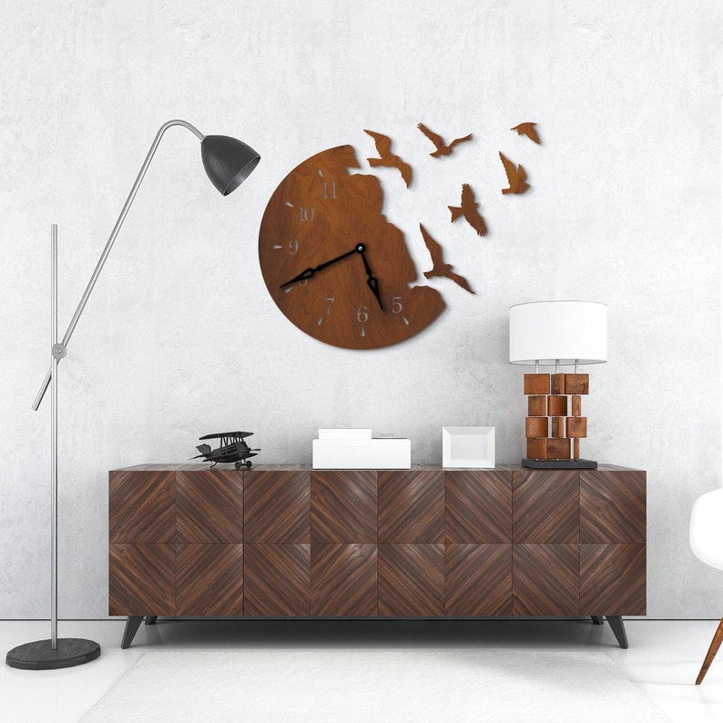 May include: A brown wooden clock with a circular face and black hands. The clock is surrounded by a design of birds flying out of the clock face. The clock is hanging on a white wall above a brown wooden cabinet.