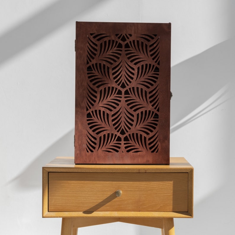 May include: A wooden cabinet with a geometric leaf pattern cut out design. The cabinet is a dark brown color and is sitting on top of a light brown wooden table with a drawer.