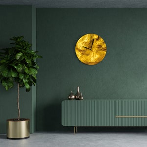 May include: A gold clock with a textured surface hangs on a dark green wall. A green cabinet with vertical lines sits on the floor in front of the clock. A potted plant with green leaves sits on the floor to the left of the cabinet.