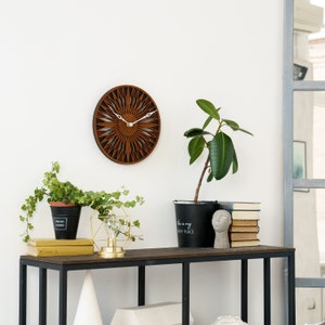 K&ouml;nnte beinhalten: Eine Wanduhr aus Holz mit Sonnenstrahlen-Design, ein schwarzes Metallregal mit einer Topfpflanze, eine kleine goldene Lampe und ein Stapel B&uuml;cher. Das Regal tr&auml;gt den Text "This is my happy place" auf der Vorderseite des Pflanzentopfs.