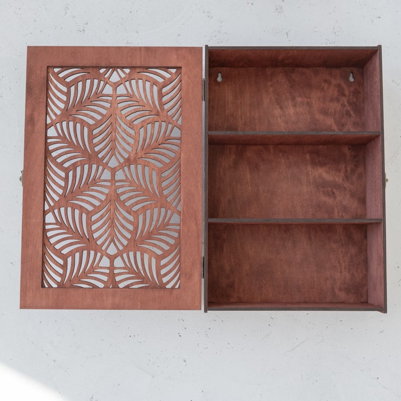 May include: A wooden cabinet with a decorative, laser-cut front panel featuring a geometric leaf pattern. The cabinet has two shelves inside and is painted a rich brown color.