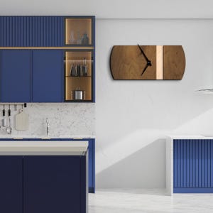 Op de afbeelding: Een moderne houten klok met een witte streep en zwarte wijzers die aan een witte muur hangt in een keuken met blauwe kasten.