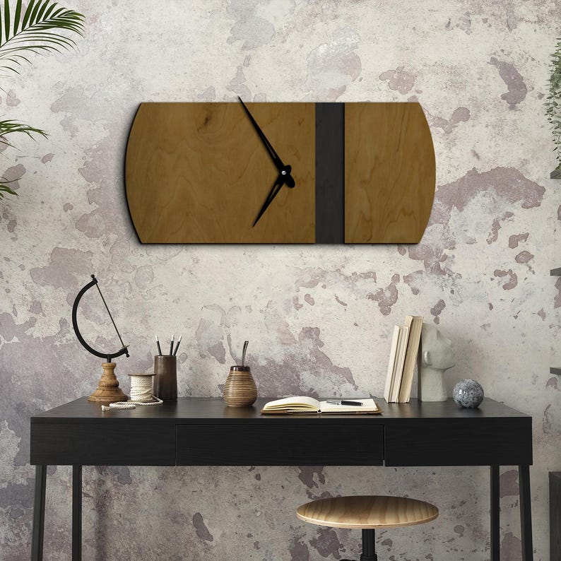 Op de afbeelding: Een houten wandklok met een zwarte metalen band in het midden. De klok is gemonteerd op een witte muur boven een zwart bureau met een boek, een vaas en een klein beeldje.