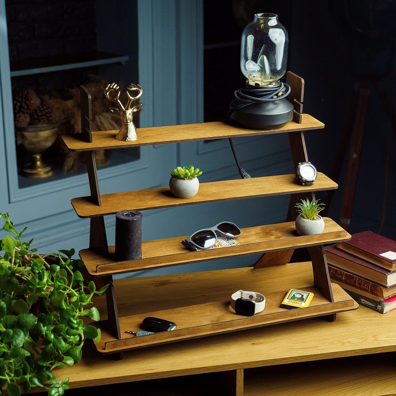 May include: A wooden tiered shelf unit with four shelves. The top shelf has a small glass lamp with a black base. The second shelf has a gold deer figurine. The third shelf has a small potted plant and a watch. The fourth shelf has a pair of sunglasses, a potted plant, and a small watch. The shelf is on a wooden table.