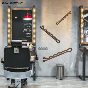 May include: Three chestnut-colored, oversized bobby pins are mounted on a gray wall. The bobby pins are wavy and decorative. The image is taken in a barber shop, with a barber chair and mirror visible in the background.