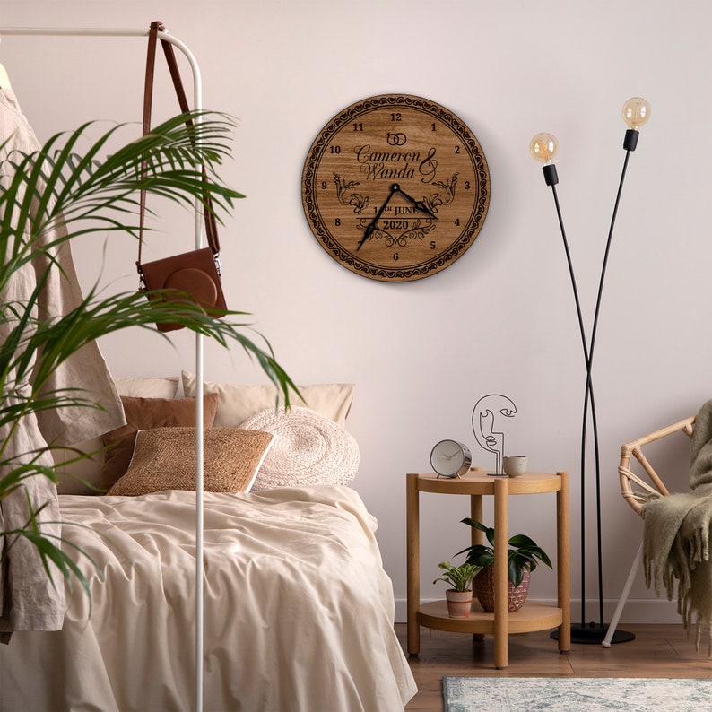 May include: A wooden clock with a brown wood grain finish. The clock face has a black border and black Roman numerals. The clock face also includes the text "Cameron & Wanda" and "June 2020". The clock is hanging on a white wall in a bedroom setting.