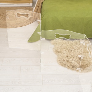 May include: A clear acrylic pet gate with bone-shaped cutouts. The gate is folded and standing on a light wood floor.