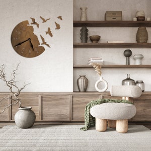 May include: A wooden wall clock with a circular design featuring a flock of birds flying out of the clock face. The clock is hanging on a white wall in a living room setting with a wooden shelf unit, a chair, and a vase with dried branches.