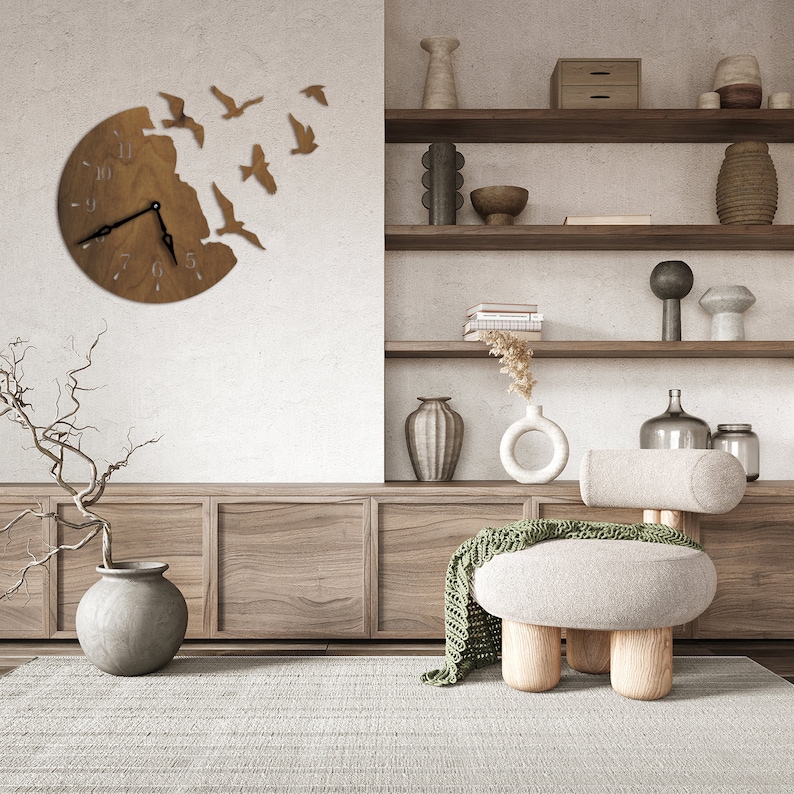 May include: A wooden wall clock with a circular design featuring a flock of birds flying out of the clock face. The clock is hanging on a white wall in a living room setting with a wooden shelf unit, a chair, and a vase with dried branches.
