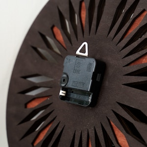 K&ouml;nnte beinhalten: Nahaufnahme der R&uuml;ckseite eines Uhrwerks mit einem schwarzen Kunststoffgeh&auml;use und einem wei&szlig;en dreieckigen Aufh&auml;nghaken. Das Uhrwerk tr&auml;gt die Aufschrift "QUARTZ" und "1988".
