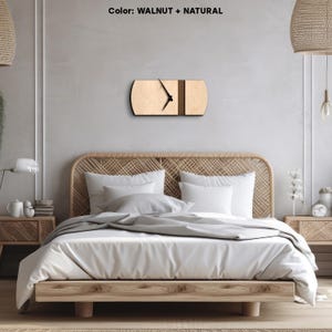 Op de afbeelding: Een houten wandklok met een natuurlijke houten afwerking en een walnootkleurige streep. De klok is gemonteerd op een witte muur boven een bed met een geweven hoofdeinde.