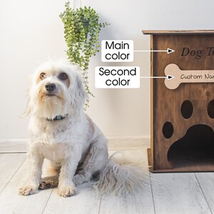 May include: A wooden dog toy storage cabinet with a paw print design and the words "Dog Toy" and "Custom Name". A small, white dog sits in front of the cabinet. The cabinet is brown and has a bone-shaped cutout.