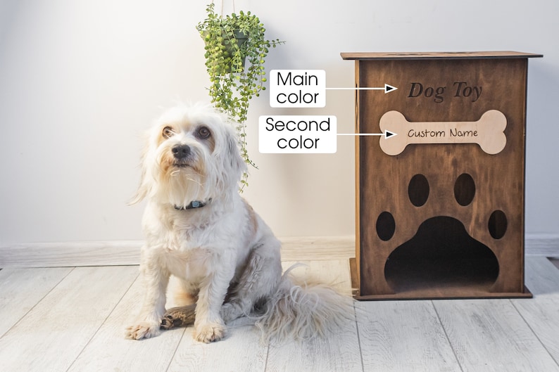 May include: A wooden dog toy storage cabinet with a paw print design and the words "Dog Toy" and "Custom Name". A small, white dog sits in front of the cabinet. The cabinet is brown and has a bone-shaped cutout.