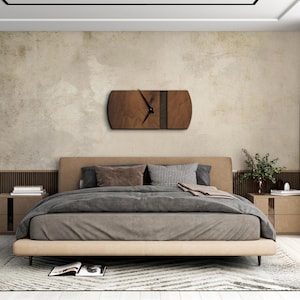 Op de afbeelding: Een houten wandklok met een donkerbruine houtnerf en een zwarte streep over de wijzerplaat. De klok hangt aan een muur in een slaapkamer met een bed en nachtkastjes.