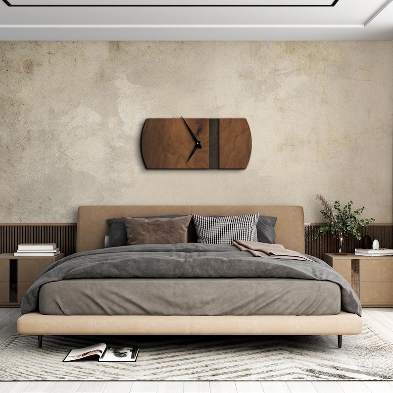 Op de afbeelding: Een houten wandklok met een donkerbruine houtnerf en een zwarte streep over de wijzerplaat. De klok hangt aan een muur in een slaapkamer met een bed en nachtkastjes.
