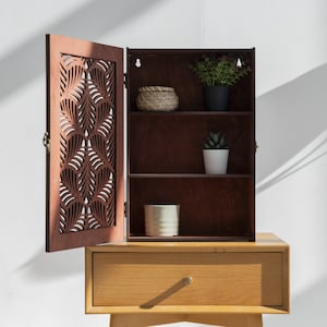 May include: A brown wooden cabinet with a decorative carved door and three shelves. The cabinet is open and has a small potted plant on the top shelf, a small potted plant on the middle shelf, and a white ceramic container on the bottom shelf.