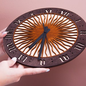 May include: A wooden clock with a sunburst design and Roman numeral markings. The clock face is brown with a white center and black hands.