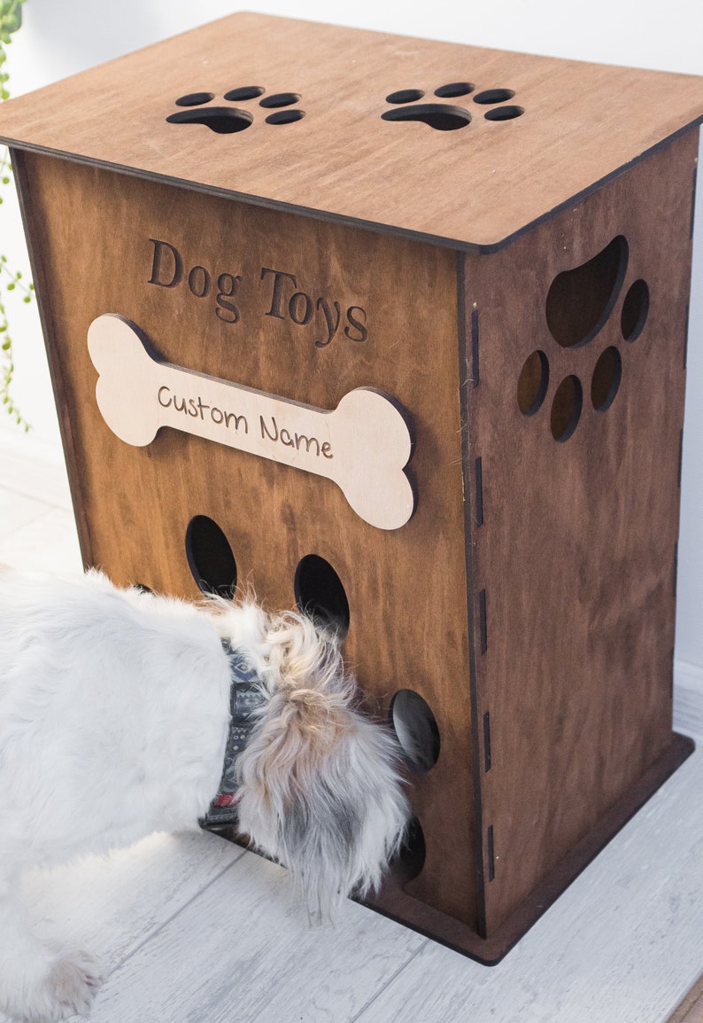May include: A wooden dog toy box with the words "Dog Toys" engraved on the front. A bone-shaped plaque below reads "Custom Name". The box has paw print cutouts and a dog is interacting with the toy.