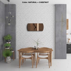 Op de afbeelding: Een houten wandklok met een natuurlijke en kastanjebruine afwerking. De klok heeft een zwarte wijzerplaat met zwarte wijzers en een witte streep in het midden. De klok hangt aan een witte bakstenen muur in een eetkamer.