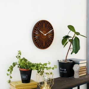 K&ouml;nnte beinhalten: Eine braune Holzuhr mit geometrischem Design und wei&szlig;en Zeigern. Die Uhr h&auml;ngt an einer wei&szlig;en Wand &uuml;ber einem Tisch mit Pflanzen und einer goldenen geometrischen Lampe.