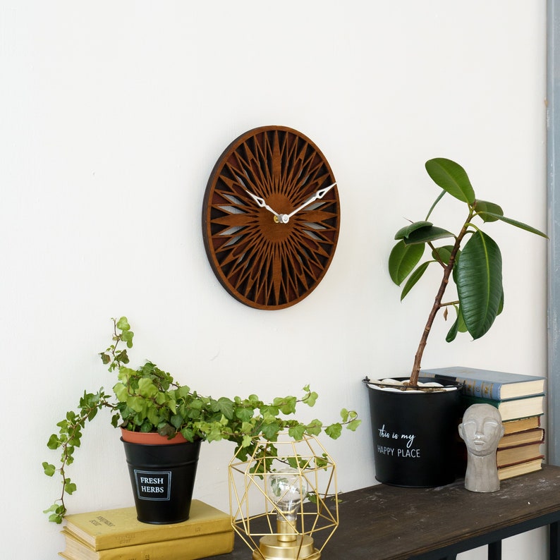 K&ouml;nnte beinhalten: Eine braune Holzuhr mit geometrischem Design und wei&szlig;en Zeigern. Die Uhr h&auml;ngt an einer wei&szlig;en Wand &uuml;ber einem Tisch mit Pflanzen und einer goldenen geometrischen Lampe.