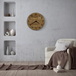 May include: A wooden clock with Roman numerals on a white wall in a living room setting. The clock is hanging above a white armchair with a brown throw blanket.