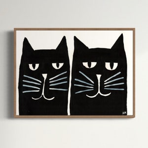 May include: A framed art print featuring two stylized black cats with white eyes and blue whiskers. The cats are painted on a white background, and the frame is a light brown color. The artwork has a minimalist, modern aesthetic.