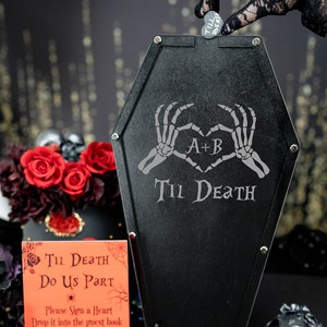 May include: Black coffin-shaped guest book alternative with the text "Til Death Do Us Part" and skeleton hands forming a heart. Includes a sign, heart-shaped tokens, and a skull. A heart token is being dropped into the coffin.
