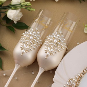 May include: Two champagne flutes decorated with pearl embellishments and gold lettering. The flutes are on a white background with white flowers.