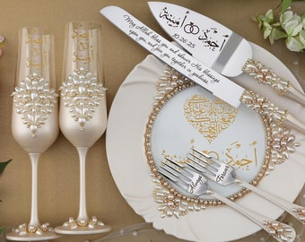 May include: A white cake server with a pearl-embellished handle and a gold inscription in Arabic script. The server is on a white plate with a gold heart design and Arabic script. Two champagne flutes with pearl embellishments and gold Arabic script are also on the plate. The inscription on the server reads "May Allah bless you and shower His blessings upon you, and join you together in goodness."