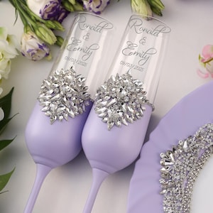 May include: Two lavender wedding toasting flutes with silver rhinestone embellishments. The flutes are engraved with the names "Ronald & Emily" and the date "09/03/2025".