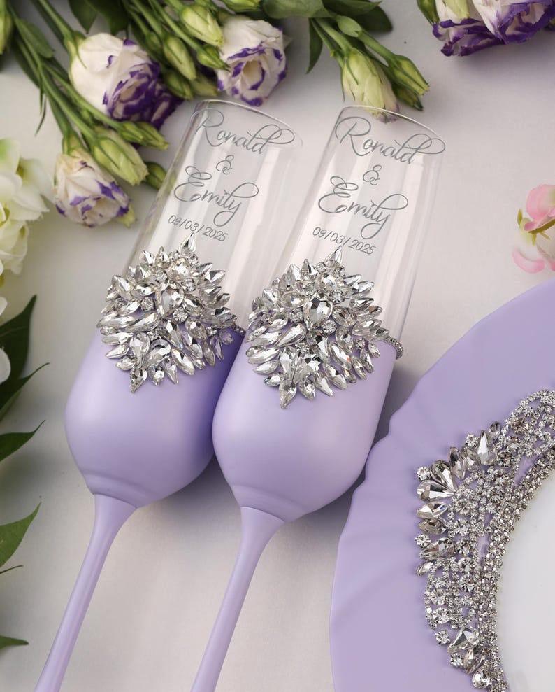 May include: Two lavender wedding toasting flutes with silver rhinestone embellishments. The flutes are engraved with the names "Ronald & Emily" and the date "09/03/2025".