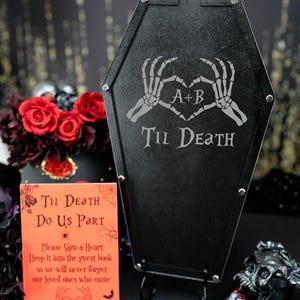 May include: A black coffin-shaped sign with the text "Til Death" and a skeleton hand heart design. The sign is on a stand with a small sign that says "Til Death Do Us Part" and a request to sign a heart and drop it into the guest book. There are black heart-shaped tokens with various phrases written on them.