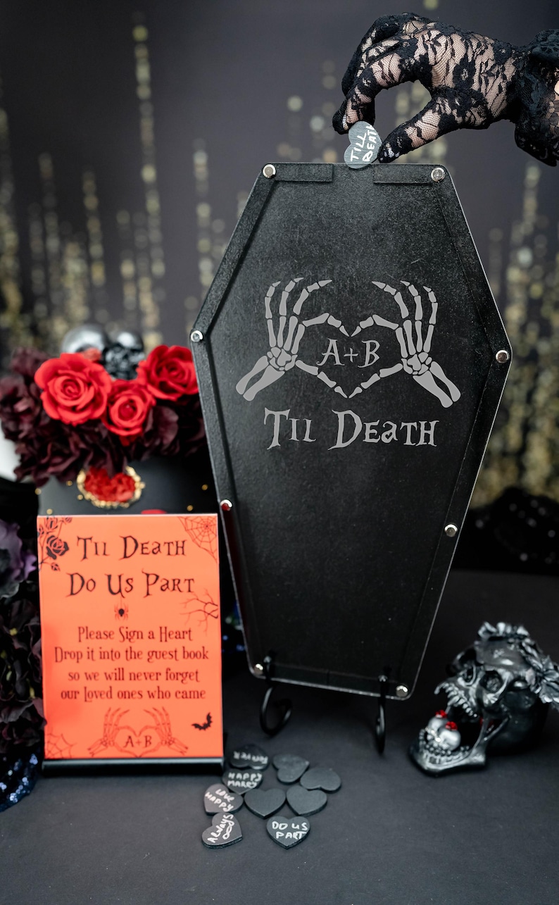 May include: A black coffin-shaped sign with the text "Til Death" and a skeleton hand heart design. The sign is on a stand with a small sign that says "Til Death Do Us Part" and a request to sign a heart and drop it into the guest book. There are black heart-shaped tokens with various phrases written on them.